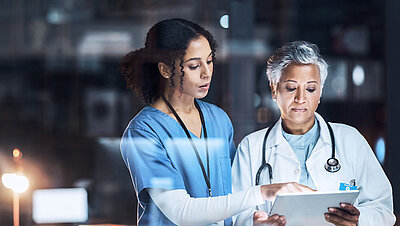 Junge Krankenschwester mit schwarzem lockigen Haar und blauem Schwesternkittel zeigt einer älteren Oberärztin mit kurzem grauen Haar in weißem Ärztinnenkittel und mit Stethoskop um den Hals etwas auf einem Tablet