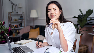 Junge brünette Frau in weißer Business-Bluse lächelt zufrieden in die Kamera und hält ihre soeben abgesetzte Brille in der Hand, während sie am Schreibtisch im Homeoffice sitzt und sich neben ihrem Laptop Notizen macht; im Hintergrund sind Pflanzen, eine Stehlampe und ein Bücherregal zu sehen